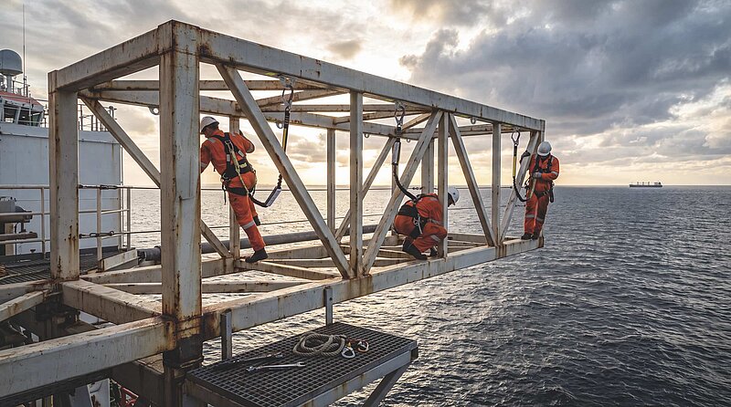 Drei Arbeiter mit Fallschutzsicherung klettern über eine Stahlkonstruktion auf einer Offshore-Plattform über dem Meer